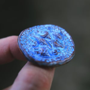 Peut inclure: Une pièce de monnaie en céramique bleue et brune avec un motif en relief de dinosaures. La pièce a une surface texturée et une apparence légèrement usée.