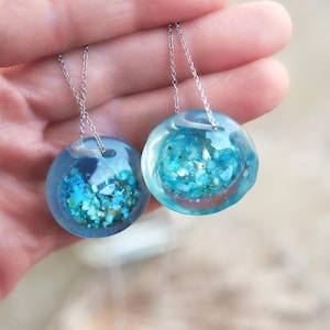 Può includere: Due collane d'argento con ciondoli rotondi in resina blu che contengono glitter turchese e dorato. I ciondoli sono di dimensioni leggermente diverse.