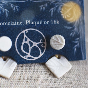 May include: A set of white porcelain earrings with leaf-like textures. The earrings include round and square shapes, displayed on a blue card with the text "Porcelaine. Plaqué or 16k". The earrings have gold-colored accents.