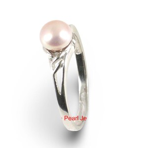 May include: A silver ring featuring a single round, pale pink pearl set at the top. The band is crafted from polished silver and displays a decorative, openwork cutout design along the shank. The text Pearl Je is visible in red at the bottom.