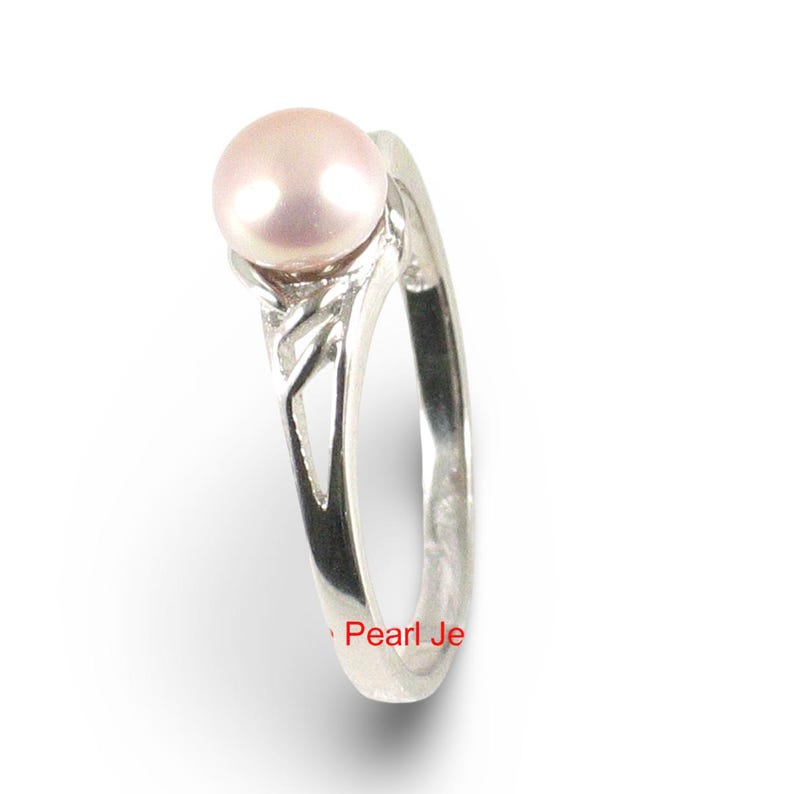 May include: A silver ring featuring a single round, pale pink pearl set at the top. The band is crafted from polished silver and displays a decorative, openwork cutout design along the shank. The text Pearl Je is visible in red at the bottom.