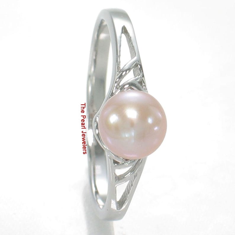 May include: A silver ring featuring a single round, pale pink pearl set in a polished metal band with an openwork, lattice-style design on the shoulders. The text The Pearl Jewelers is printed vertically along the side of the ring band.