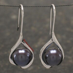 May include: A pair of silver-toned drop earrings, each featuring a large, dark gray pearl. The pearls are cradled within a stylized, teardrop-shaped setting. The earrings hang from a simple hook. The Pearl Jewelers is printed on the side.