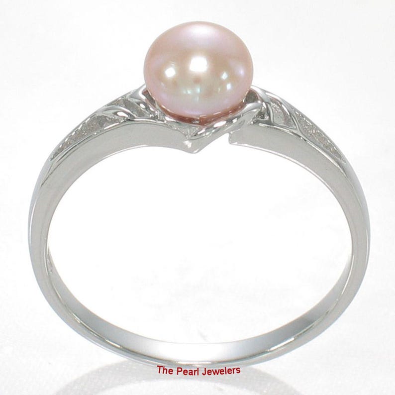 May include: A silver-toned ring featuring a single round, pale pink pearl set at the center. The band has a polished finish with textured, leaf-like detailing on the shoulders leading up to the pearl setting. The text The Pearl Jewelers is visible.