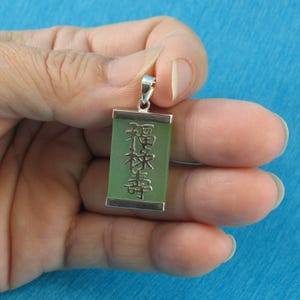 May include: A rectangular jade pendant with silver-colored metal trim. The green jade has engraved Chinese characters. The pendant is suspended from a silver-colored bail. The background is a solid light blue.