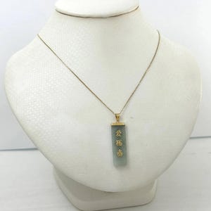 May include: A gold-toned necklace with a rectangular pendant. The pendant is a light green color with gold lettering. The necklace is displayed on a white mannequin bust.