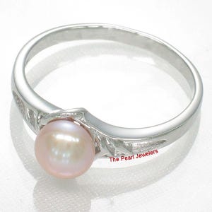 May include: A silver ring featuring a single round, light pink pearl set in the center. The band has a polished finish with decorative cut-out details on the sides. The text The Pearl Jewelers is printed in red near the base of the setting.