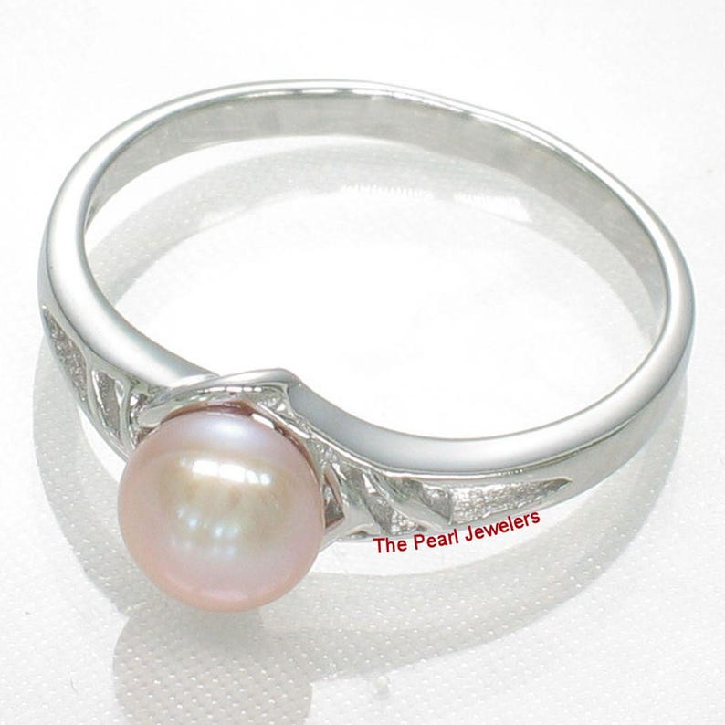 May include: A silver ring featuring a single round, light pink pearl set in the center. The band has a polished finish with decorative cut-out details on the sides. The text The Pearl Jewelers is printed in red near the base of the setting.
