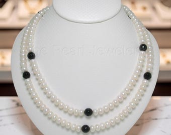 Puede incluir: Un collar de doble hebra con perlas blancas intercaladas con cuentas negras lisas. El collar se muestra sobre un busto blanco, con el texto The Pearl Jeweler visible al fondo. El diseño consiste en perlas blancas y cuentas negras alternadas.