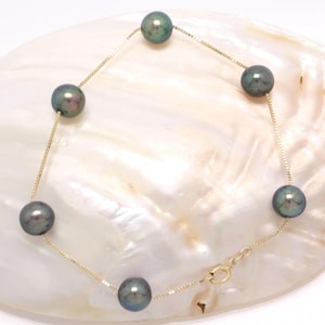 May include: A gold chain bracelet featuring six dark gray pearls. The pearls are evenly spaced along the delicate chain, which fastens with a small clasp. The bracelet is displayed on a white, textured surface.