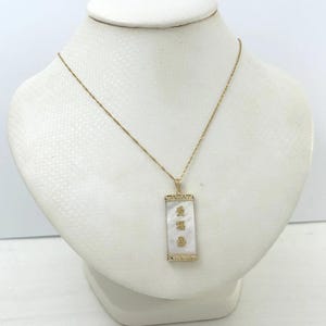 May include: A gold-colored necklace with a rectangular pendant. The pendant has a white, mother-of-pearl inlay with gold-colored Chinese characters. The necklace is displayed on a white bust.