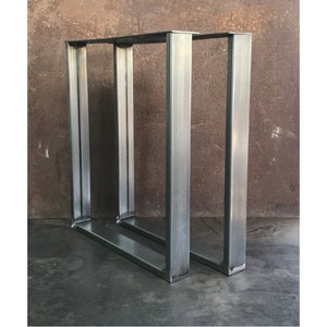 May include: Two silver metal table legs with a square frame design. The legs are made of thick metal and have a brushed finish.