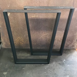 May include: Two black metal table legs, each with a rectangular frame, are shown on a gray concrete floor. The legs are made of thick, sturdy metal and have a simple, modern design.