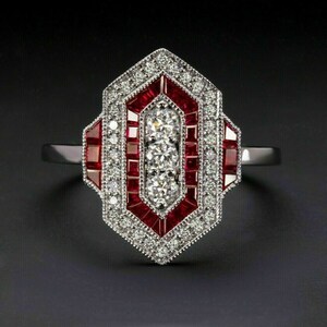 May include: A silver ring with a geometric design. The ring features a central row of three round diamonds, surrounded by red gemstones and smaller white diamonds. The ring is set against a black background.