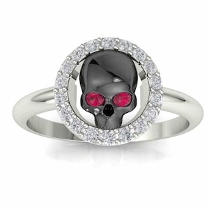May include: A silver ring with a black skull design and red gem eyes. The skull is surrounded by a halo of small white gems.