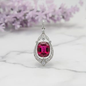 May include: A silver pendant with a large, rectangular, deep pink gemstone at its center. The gemstone is surrounded by a halo of smaller, clear gemstones. The pendant has an ornate, Art Deco-inspired design.