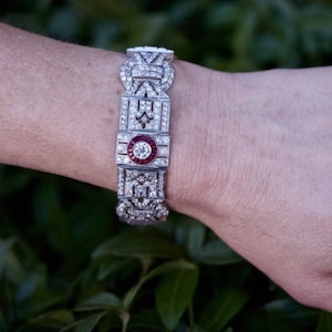 May include: A silver bracelet with a geometric design and a large red ruby in the center. The bracelet is set with diamonds.
