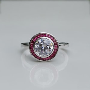 May include: A silver ring featuring a large, round, clear gemstone at the center, surrounded by a halo of small, rectangular, red gemstones. The ring's band is thin and polished, reflecting the light. The design is elegant and classic.