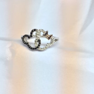 May include: A silver ring with two interlocking hearts, one with black stones and the other with white stones. A small bow is attached to the top of the ring.