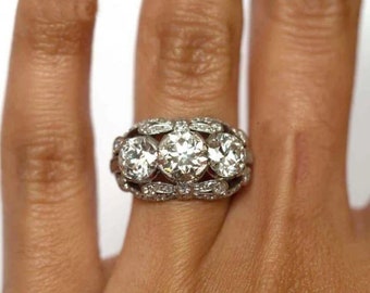 Könnte beinhalten: Ein silberner Ring mit drei großen runden Diamanten, die in einem dekorativen Band gefasst sind. Die Diamanten sind von kleineren Diamanten umgeben.