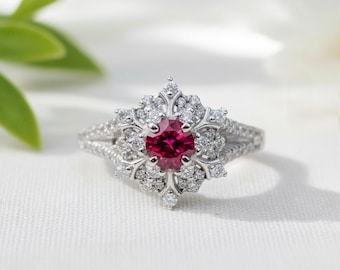 May include: A silver ring with a central round, deep red gemstone, surrounded by a halo of smaller, clear gemstones. The ring's band splits into two, creating a delicate, open design. The ring is set against a white background.