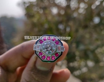 May include: A silver ring with a large round clear stone surrounded by smaller pink stones. The ring has a black and green enamel design.