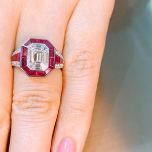 May include: A rectangular-cut diamond ring with a red ruby halo and accents. The ring features a large, clear center stone surrounded by smaller red rubies and diamond accents on the band. The ring is set in a silver or white gold setting.