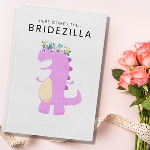 May include: A white journal with the text "HERE COMES THE... BRIDEZILLA" and a cartoon dinosaur wearing a floral crown. A bouquet of pink roses and a lace ribbon are on a pink background.