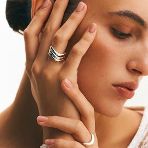 Può includere: Una mano di donna con due anelli d'argento. L'anello sull'indice è una fascia impilata con tre fasce curve. L'anello sul medio è una singola fascia curva.