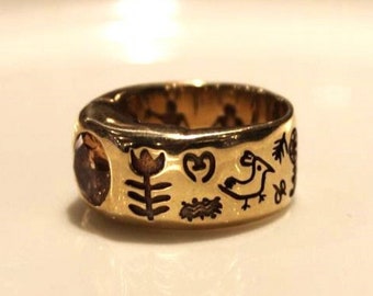 May include: A gold ring with a dark brown gemstone and engraved symbols, including a bird, a heart, a flower, and a tree.