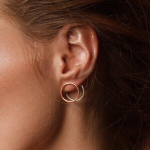 May include: A pair of small, delicate earrings with two interlocking circles. The outer circle is a warm rose gold color, and the inner circle is a cool silver color.