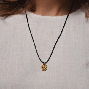 May include: A gold pendant necklace with a detailed design hangs from a black cord. The necklace is worn by a person with long brown hair.