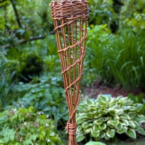 Könnte beinhalten: Eine handgefertigte, braune Weidenholzfackel, ideal für die Außenbeleuchtung. Die Fackel hat eine konische Form mit einem eng gewebten Oberteil und einem spiralförmigen Geflecht am Körper. Die Basis ist gebündelt und gebunden. Der Hintergrund ist ein üppiger grüner Garten.
