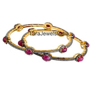 May include: Two gold bangles featuring oval ruby gemstones and small diamond accents. The bangles have a dark grey band with gold detailing. The text "TaraJewelsIndia" is visible. The bangles are displayed on a white background.