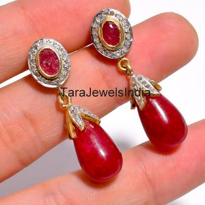 Peut inclure: Boucles d'oreilles pendantes avec des pierres précieuses ovales rouge foncé suspendues à des montures dorées. Les boucles d'oreilles ont un halo argenté de petites pierres autour des pierres rouges. Le texte "TaraJewelsIndia" est visible.