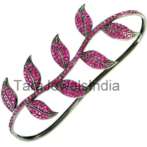 May include: A dark metal two-finger ring featuring a branch design with six leaves. Each leaf is encrusted with small, vibrant pink stones. The text "TaraJewelsIndia" is visible across the center of the ring, which has an elongated oval shape.