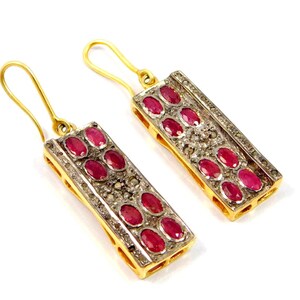 May include: A pair of gold-colored rectangular earrings with a row of oval-shaped red gemstones. The earrings are accented with small, clear gemstones and have gold-colored hooks.