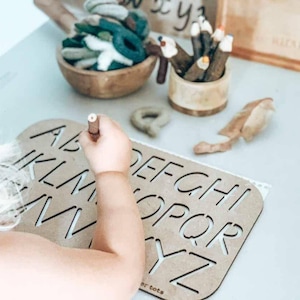 May include: A wooden alphabet tracing board with the letters A through Z cut out. A child is tracing the letters with a wooden stylus.