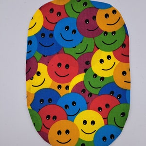 May include: A colourful oval fabric patch with a repeating pattern of smiling faces in various colours, including yellow, red, blue, green, and purple.