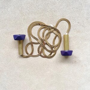 Op de afbeelding: Een goudkleurige visgraatketting met twee bijpassende paarse en goudkleurige geometrische hangers. De ketting is in een luspatroon op een neutrale achtergrond gerangschikt.