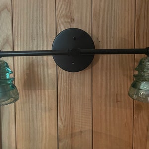May include: A wall-mounted light fixture featuring a matte black circular base and a horizontal metal arm. Two light sockets are fitted with vintage-style, translucent light green glass insulators that serve as decorative shades for the bulbs.