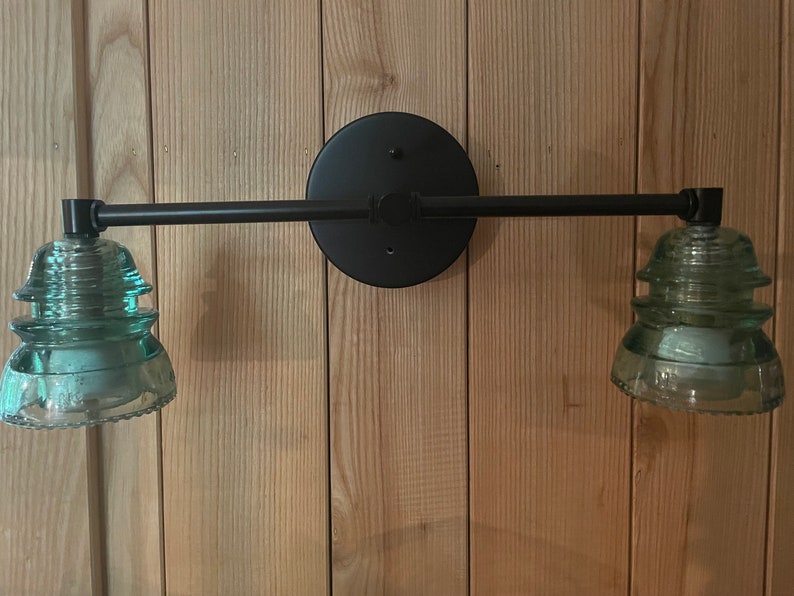 May include: A wall-mounted light fixture featuring a matte black circular base and a horizontal metal arm. Two light sockets are fitted with vintage-style, translucent light green glass insulators that serve as decorative shades for the bulbs.