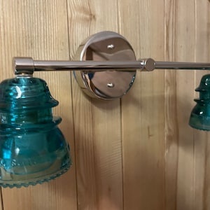 May include: A wall-mounted light fixture featuring a polished chrome horizontal bar and a circular base plate. Two teal-colored glass insulators serve as shades, each with a stepped, tiered profile and embossed lettering along the bottom edge.