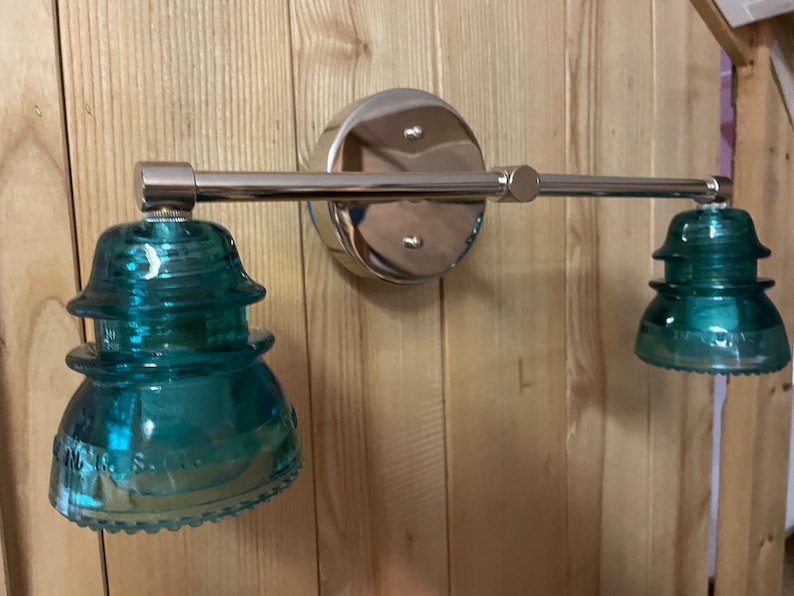 May include: A wall-mounted light fixture featuring a polished chrome horizontal bar and a circular base plate. Two teal-colored glass insulators serve as shades, each with a stepped, tiered profile and embossed lettering along the bottom edge.