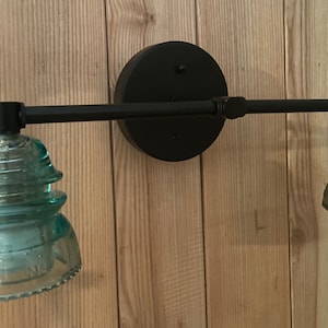 May include: A wall-mounted light fixture featuring a matte black horizontal metal bar and a circular backplate. Two light sockets are fitted with translucent, pale teal glass insulators that have a tiered, stepped design and a wide base.