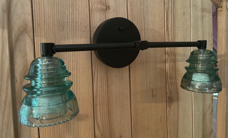 May include: A wall-mounted light fixture featuring a matte black horizontal metal bar and a circular backplate. Two light sockets are fitted with translucent, pale teal glass insulators that have a tiered, stepped design and a wide base.