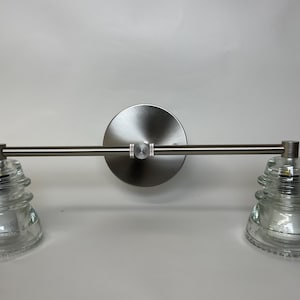 May include: A wall-mounted light fixture featuring a brushed nickel horizontal bar and a circular backplate. Two clear, tiered glass insulators are attached to the ends of the bar, serving as the light shades for this dual-bulb lighting unit.