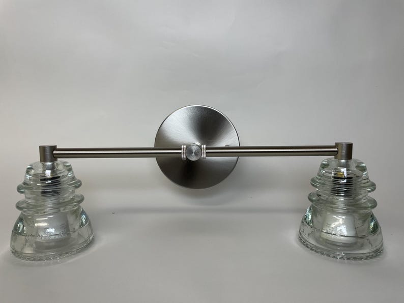 May include: A wall-mounted light fixture featuring a brushed nickel horizontal bar and a circular backplate. Two clear, tiered glass insulators are attached to the ends of the bar, serving as the light shades for this dual-bulb lighting unit.