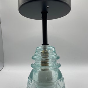 May include: A black metal ceiling fixture with a clear glass insulator pendant. The insulator is made of three stacked glass pieces with a textured surface. The fixture is designed for a single light bulb.