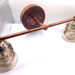 May include: A wall-mounted light fixture featuring a horizontal copper-toned metal bar attached to a circular backplate. Two clear, tiered glass insulators are secured to the ends of the bar, serving as the light shades for this industrial-style lamp.
